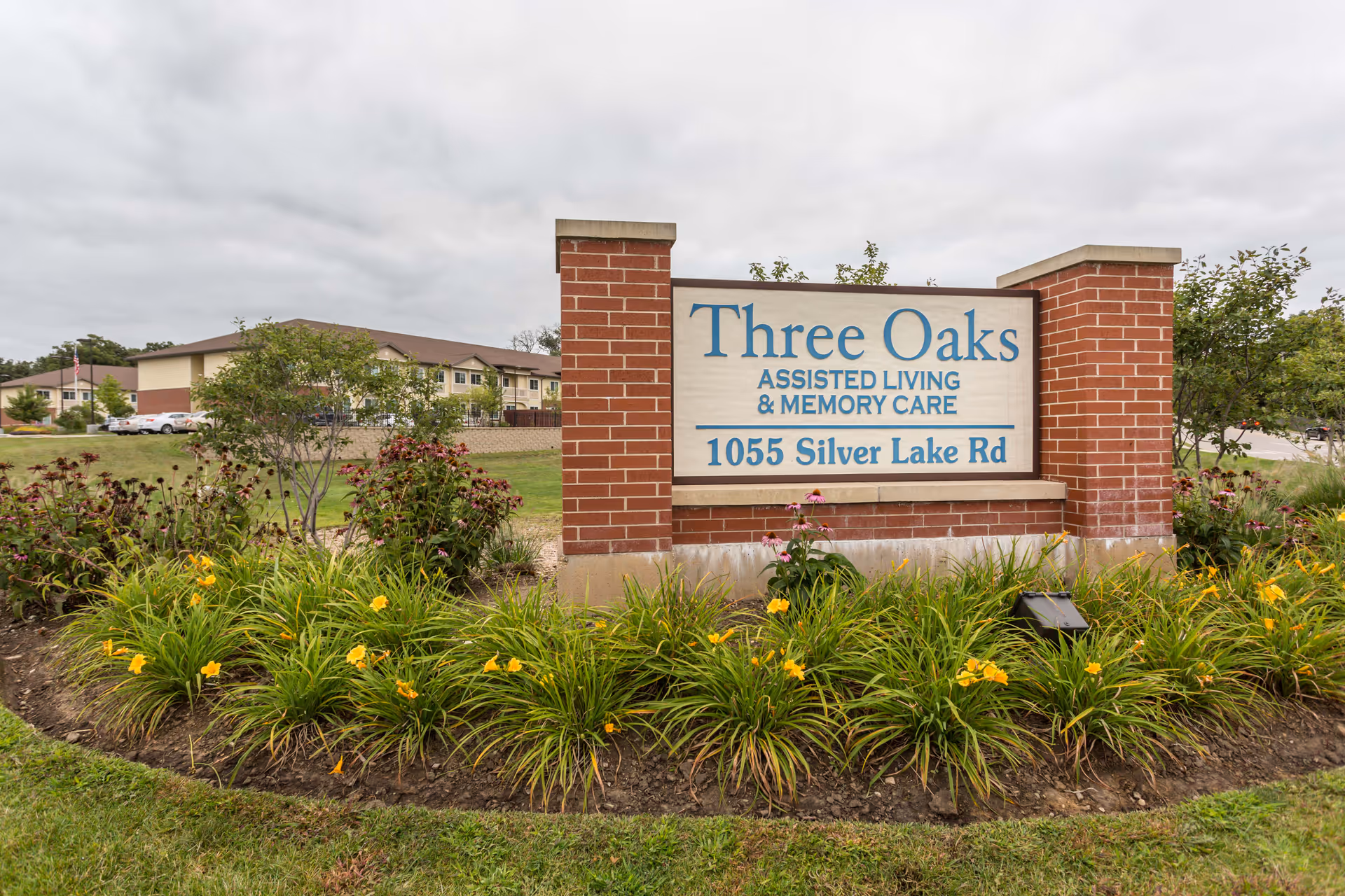 A brick sign for Three Oaks Assisted Living & Memory Care at 1055 Silver Lake Rd, surrounded by green plants and yellow flowers, with the facility building and parked cars visible in the background under a cloudy sky.