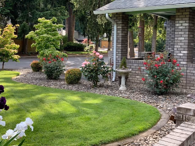 A well-maintained garden area with green grass, flowering bushes, and small trees next to a brick building with a covered porch. There is a birdbath among the plants and a stone pathway bordering the grass.