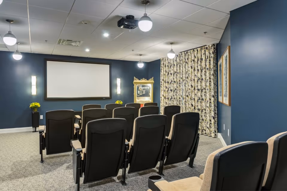 A small theater room with two rows of black and beige cushioned chairs facing a large blank projection screen on a dark blue wall. There are three round ceiling lights and two wall sconces providing illumination. A popcorn machine is visible in the corner next to patterned curtains, and two framed pictures hang on the right wall.