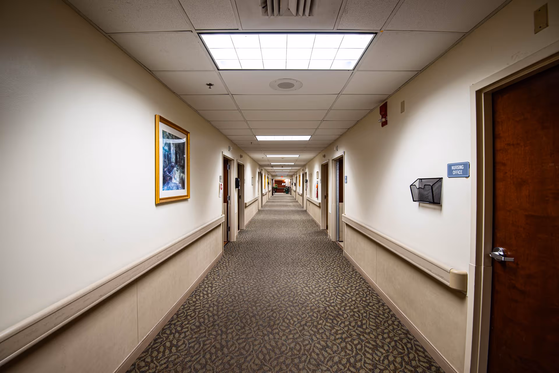 A long, well-lit hallway in a nursing and rehabilitation center with beige walls, patterned carpet, and several closed doors on both sides. There is a framed picture on the left wall and a sign labeled 'Nursing Office' on the right wall near a wooden door.