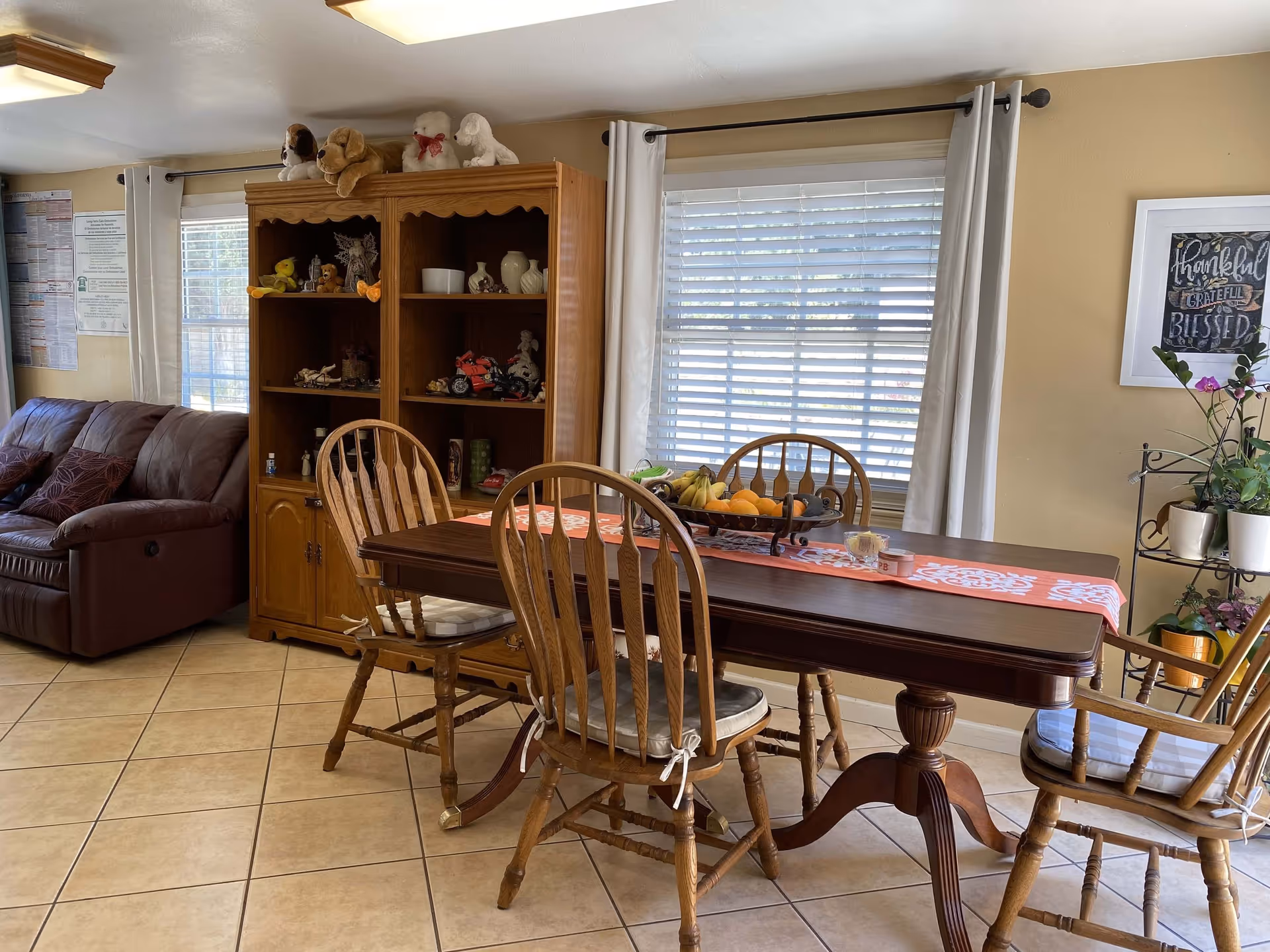 A cozy dining area with a wooden dining table and four wooden chairs with cushions. The table has a decorative runner and a fruit bowl with bananas, oranges, and other fruits. Behind the table is a large window with white blinds and white curtains. To the left, there is a wooden cabinet filled with various decorative items and stuffed animals on top. A brown leather couch is partially visible on the far left. The walls are painted beige, and there is a framed artwork on the right wall with the words 'thankful, grateful, blessed'. The floor is tiled with light-colored tiles.