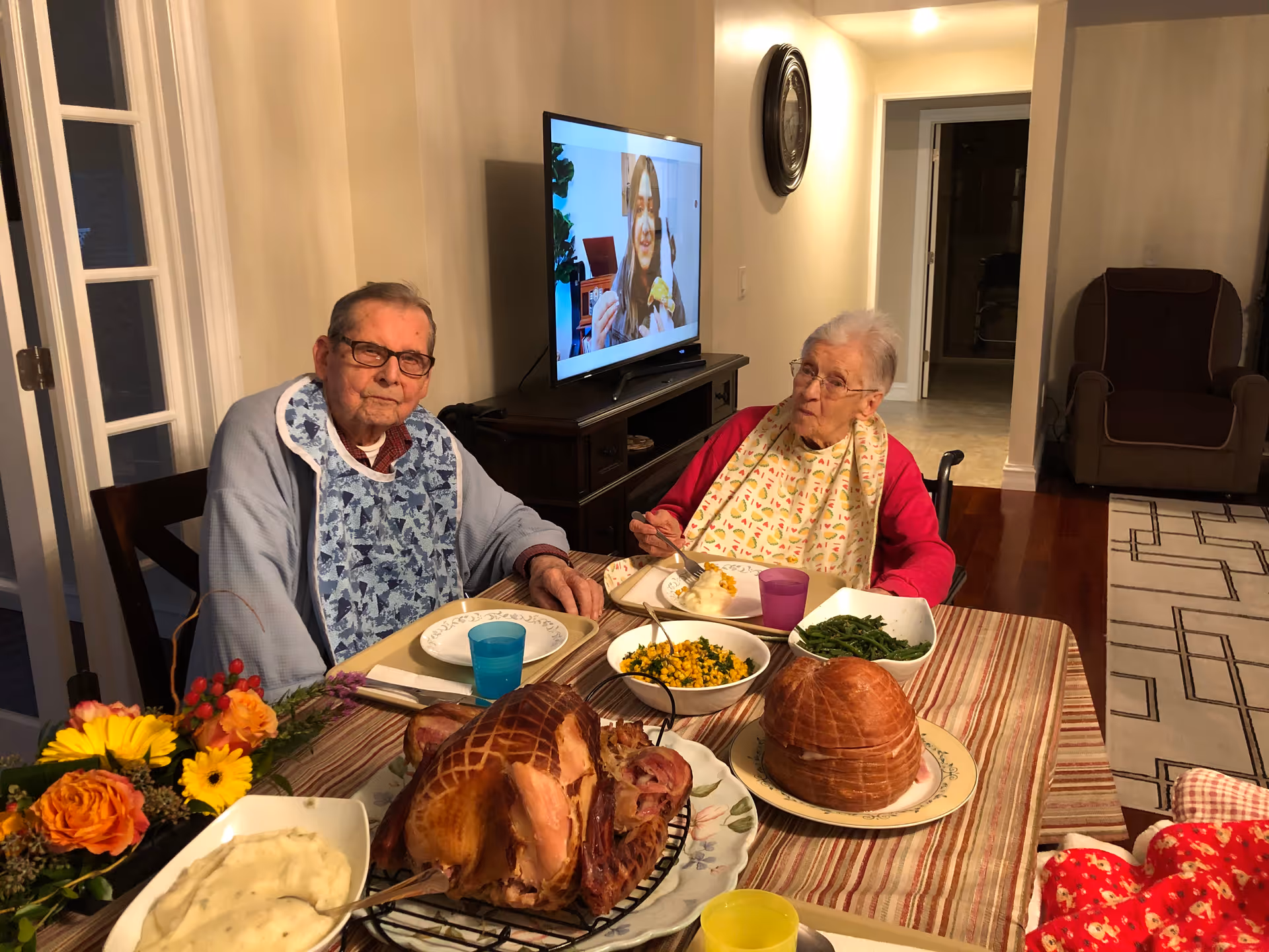 Two elderly individuals sitting at a dining table with a meal in front of them, including a large cooked ham, mashed potatoes, green beans, and corn. Both are wearing bibs and appear to be in a cozy dining area with a television in the background showing a woman eating. There are flowers on the table and a comfortable chair in the background.