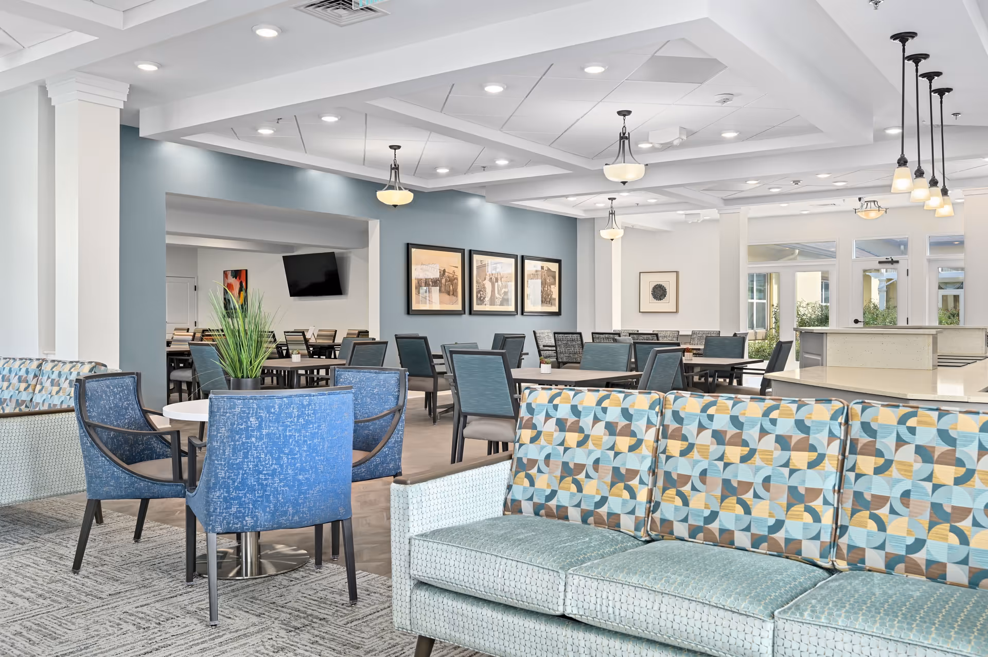 A bright and spacious common area in a senior living facility featuring patterned sofas and blue upholstered chairs around small round tables. The room has a light blue accent wall with framed artwork, multiple ceiling lights, and large windows letting in natural light. In the background, there are more tables and chairs arranged for dining or socializing.