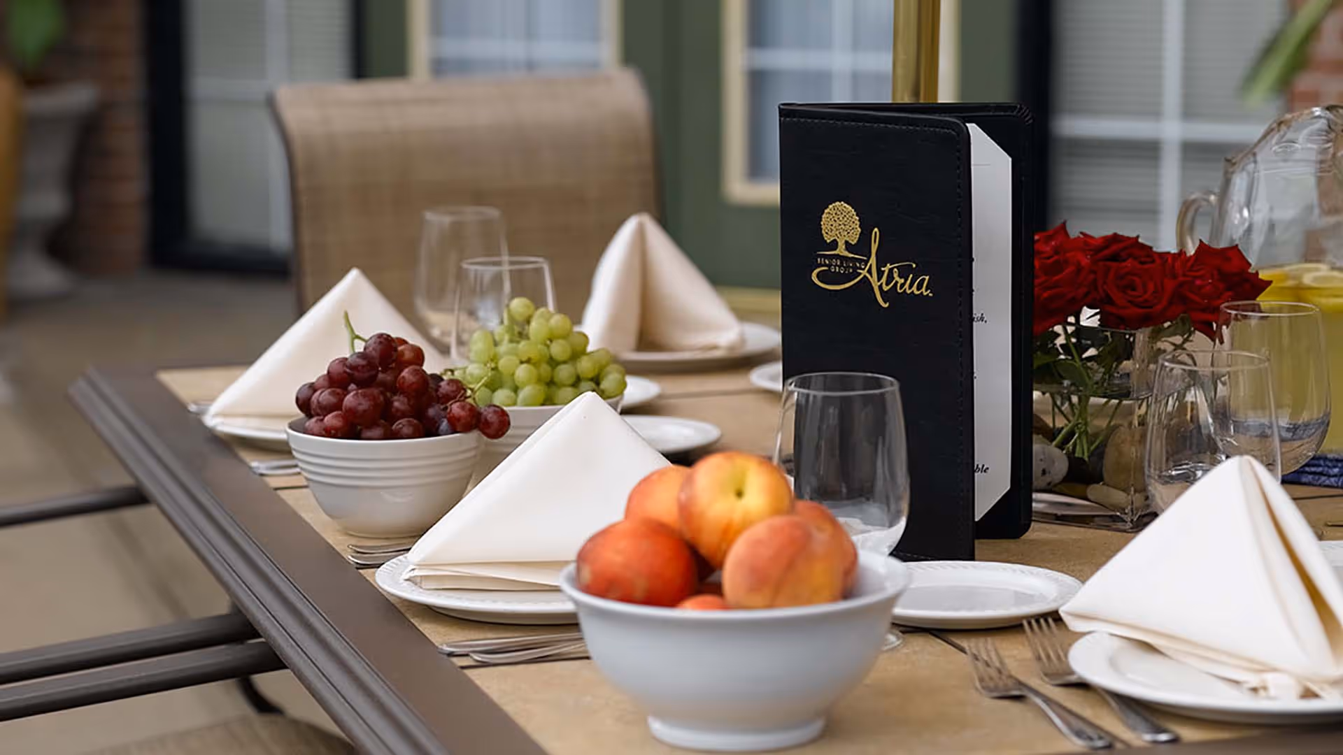 A dining table set with white plates, folded white napkins, clear drinking glasses, bowls of fresh fruit including peaches and grapes, a black menu with gold lettering reading 'Atria', and a vase of red roses in the background.