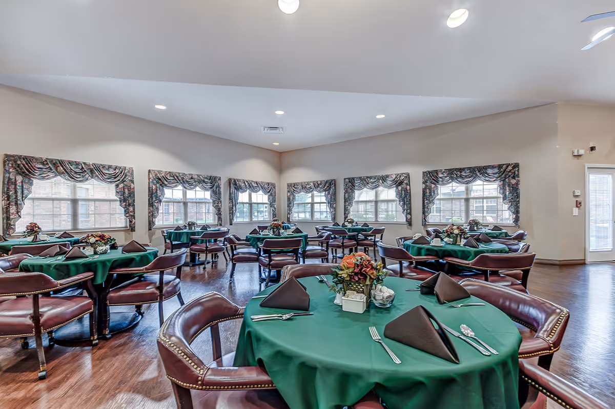 Bright dining room with multiple round tables covered in green tablecloths, leather chairs, and floral centerpieces beneath large windows.