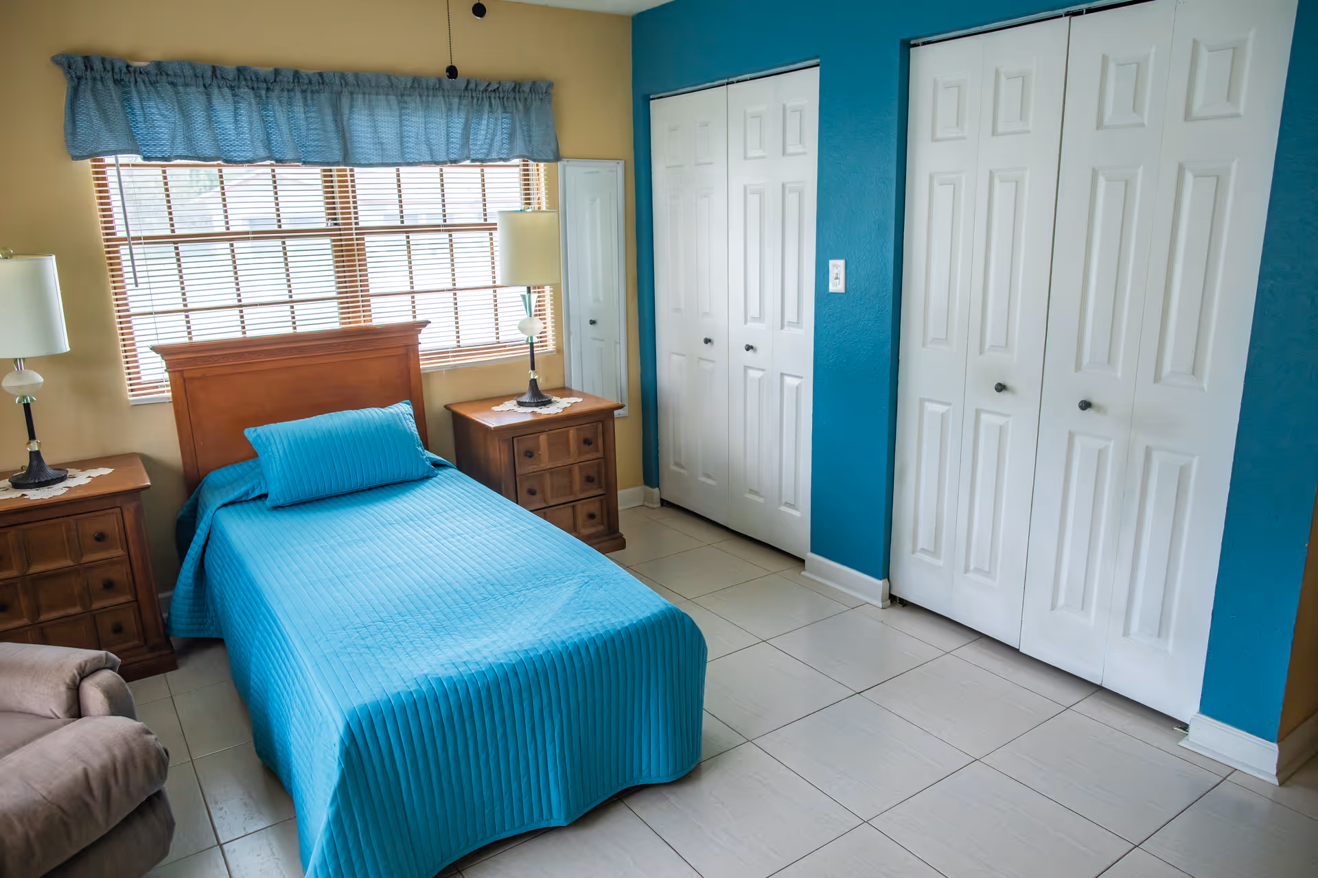 A bedroom with a single bed covered in a blue bedspread and matching pillow. The room has two wooden nightstands with lamps on each side of the bed, a large window with blinds and a blue valance, a beige armchair, and two white closets with paneled doors against a blue accent wall.
