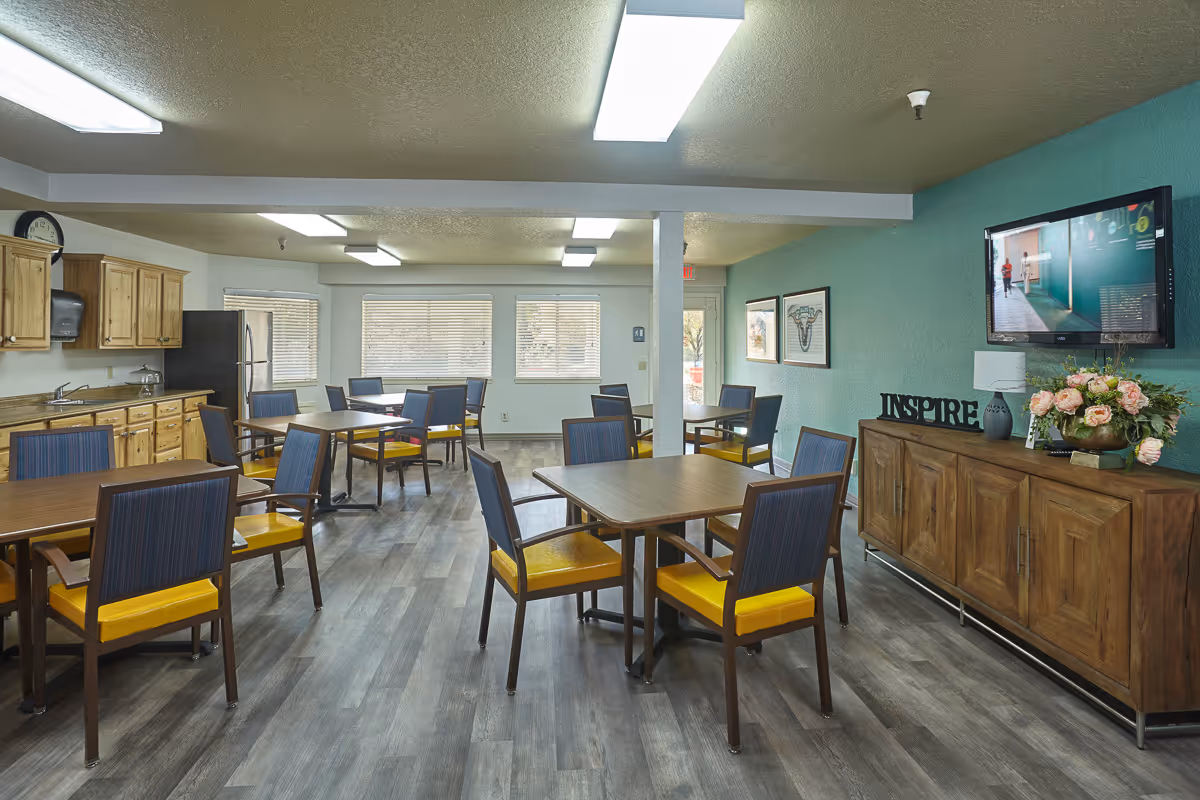 A bright dining room with several square wooden tables and chairs with yellow seats and blue backs. The room has wood flooring, a kitchen area with wooden cabinets, a refrigerator, and a sink on the left side. On the right wall, there is a mounted flat-screen TV above a wooden sideboard decorated with a flower arrangement and a decorative sign that says 'INSPIRE'. Large windows at the back let in natural light.