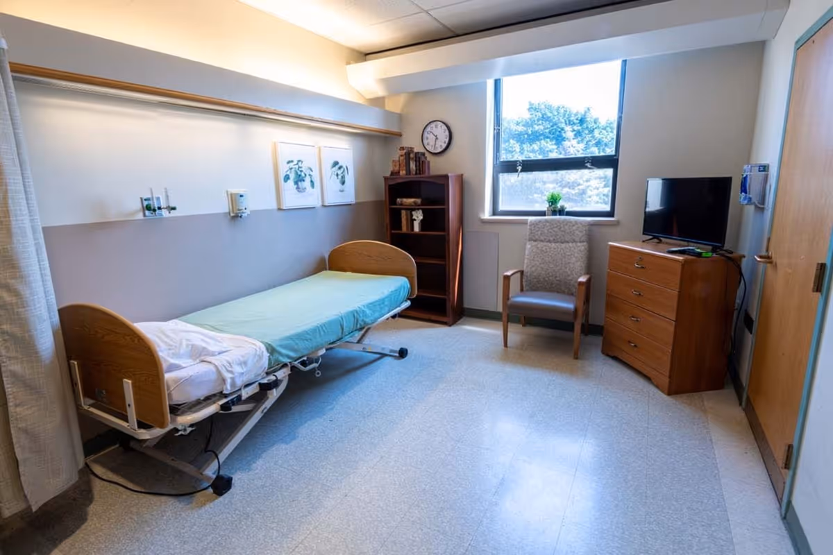 A simple senior living bedroom with an adjustable single bed, armchair, dresser with TV, bookshelf and a window.