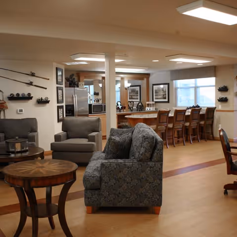 A cozy living area with patterned armchairs and a round wooden table in the foreground. In the background, there is a kitchen area with a refrigerator, microwave, and a counter with several wooden bar stools. The room has wooden flooring and soft lighting from ceiling fixtures.