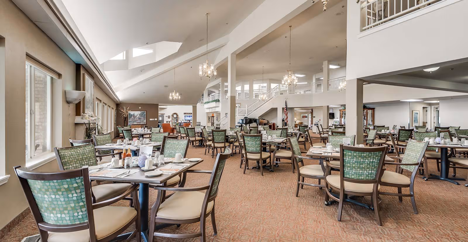 Spacious dining room with multiple tables and chairs arranged neatly. Each table is set with cups, plates, and condiments. Large windows on the left side allow natural light to fill the room. The ceiling is high with chandeliers hanging down, and there is a staircase and balcony area visible in the background.