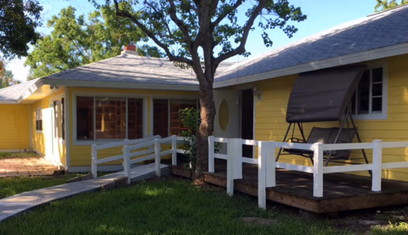 Exterior view of a yellow single-story building with a white railing and a wooden deck featuring a swing seat. There is a tree in front of the building and a concrete pathway leading to the entrance. The area is surrounded by green grass and trees.