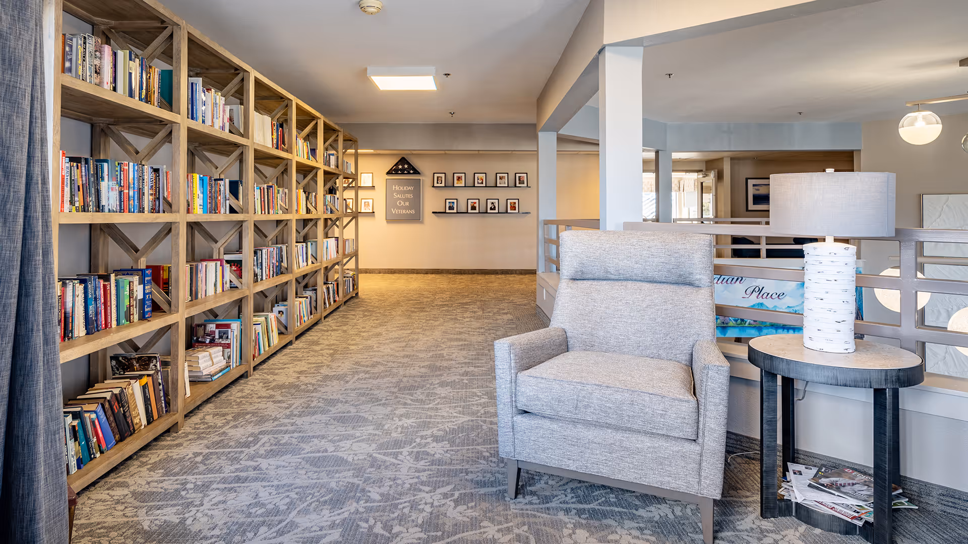 A cozy reading nook in a senior living facility featuring a large wooden bookshelf filled with books along the left wall, a comfortable gray armchair, and a small round side table with a lamp. The area has carpeted flooring and soft lighting, with a hallway leading to a wall displaying framed photos and a sign that reads 'Holiday Salutes Our Veterans.'