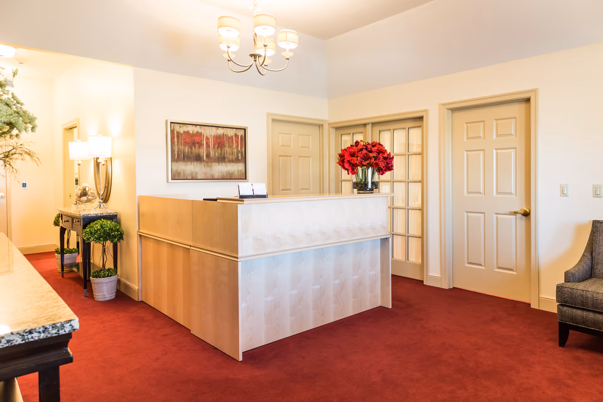 Reception area with a light wood reception desk adorned with a vase of red flowers. The room has beige walls, red carpet, and several closed doors including one with glass panels. A chandelier with multiple lights hangs from the ceiling. There is a small table with a lamp and decorative plants on the left side and a gray upholstered chair on the right.