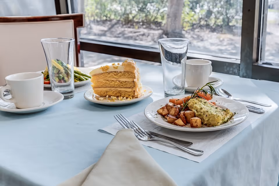 Table set near a window with a plated entrée of potatoes and fish, a slice of layered cake, cups and glasses on a light blue tablecloth.