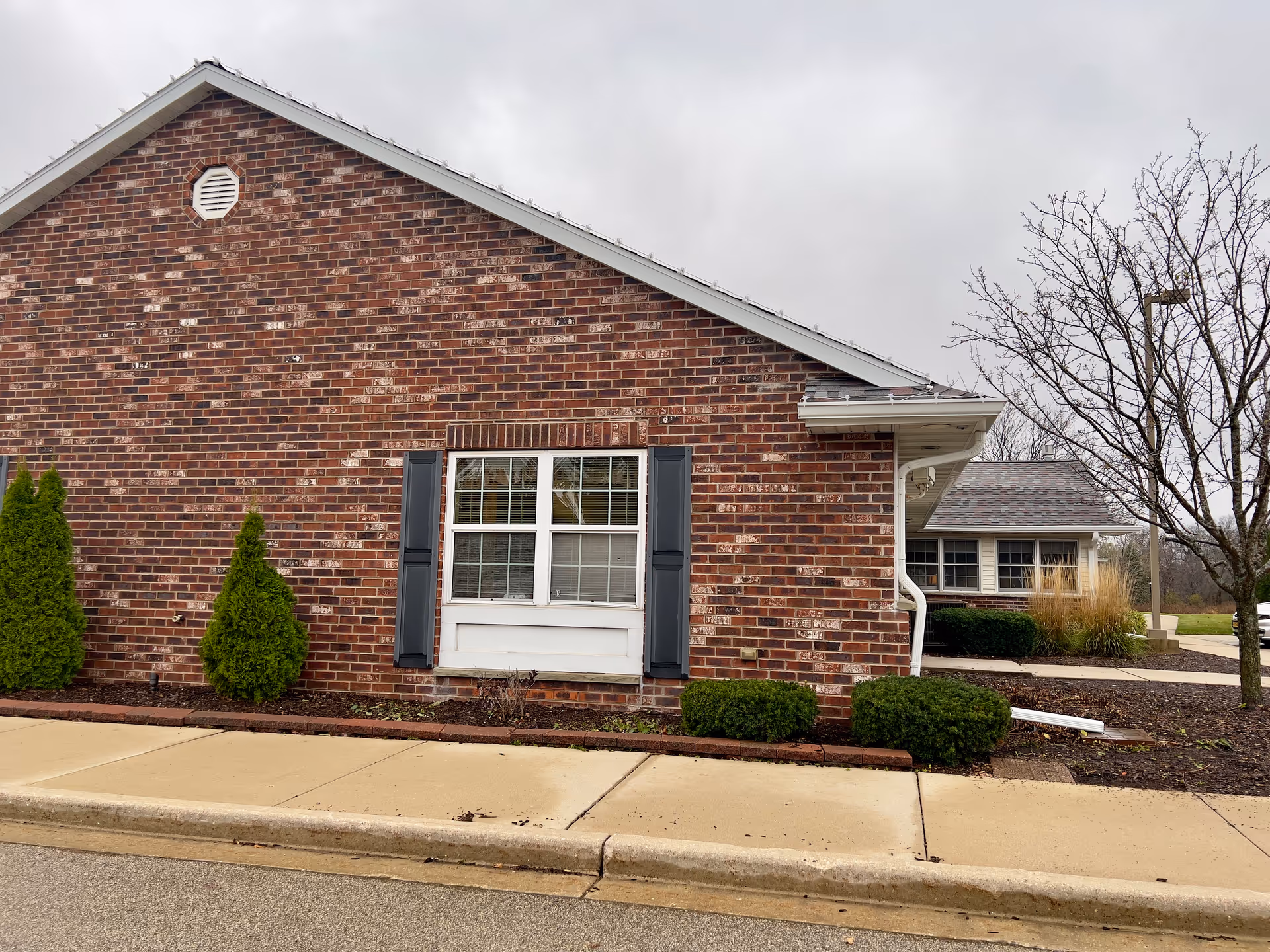 Brick exterior wall of a single-story building with a window, shrubs, sidewalk, and a leafless tree under a cloudy sky.
