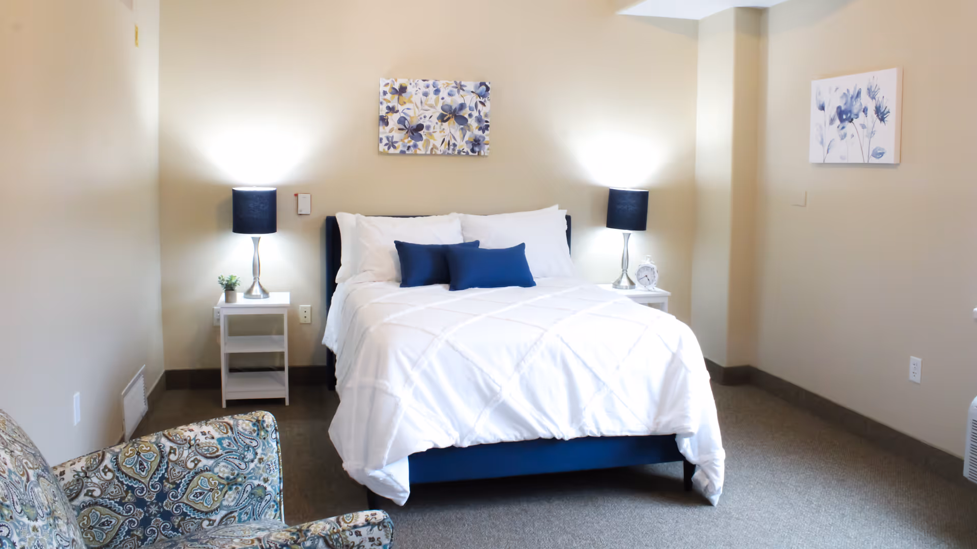 Well-lit bedroom with a neatly made bed, two nightstands and lamps, wall art, and a patterned armchair.