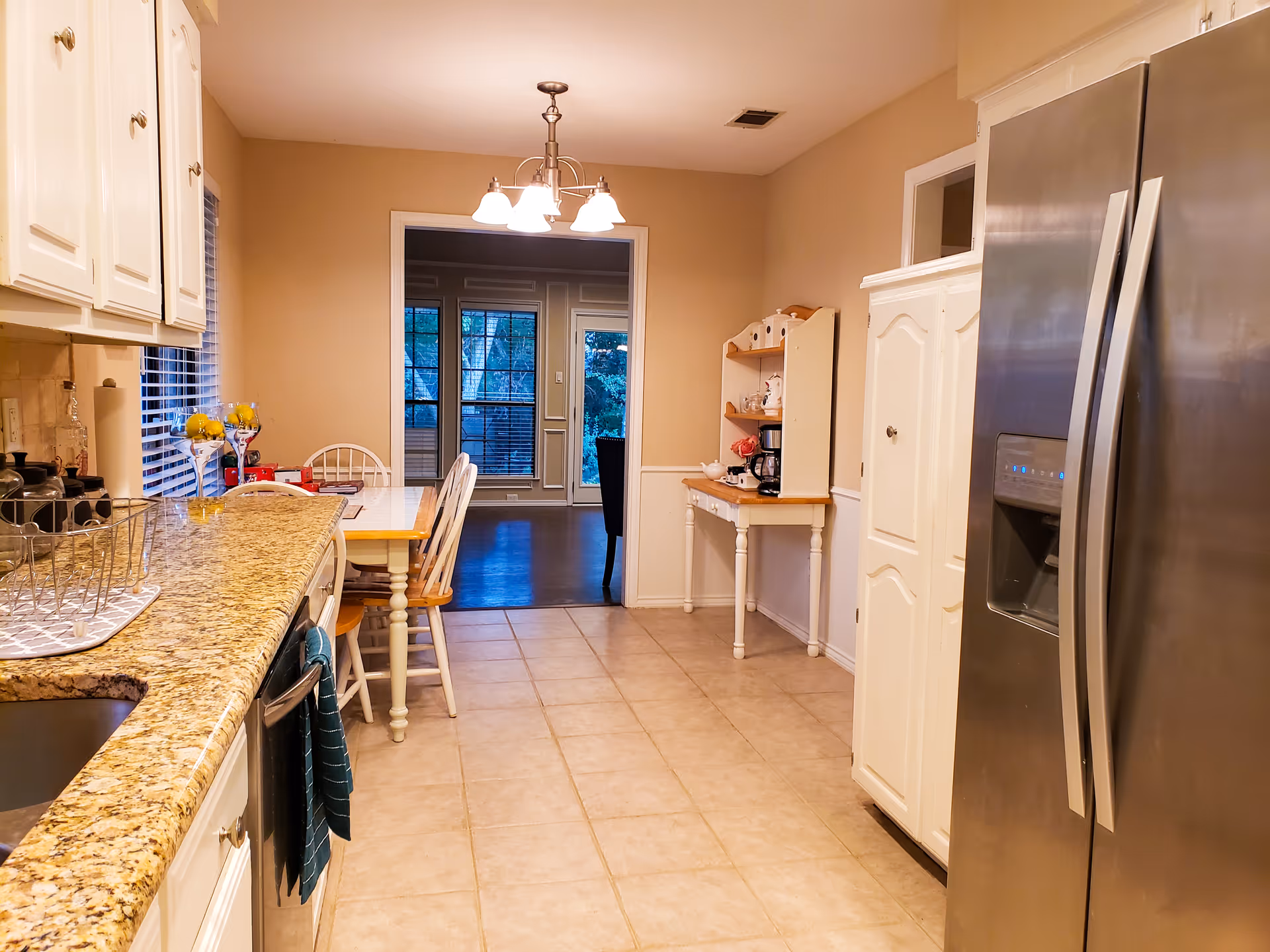 A bright kitchen with granite countertops, white cabinets, a stainless steel refrigerator, and a small dining table with chairs. There is a hanging light fixture above the table and a window with blinds on the left side. The kitchen opens into another room with large windows and a door leading outside.