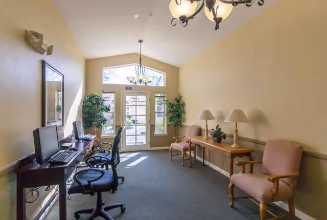 A bright room with beige walls and carpeted floor featuring a row of computer desks with chairs on the left side and a wooden table with two lamps and two upholstered chairs on the right side. Large windows and a glass door at the far end let in natural light, with two potted plants flanking the door. A chandelier hangs from the ceiling.