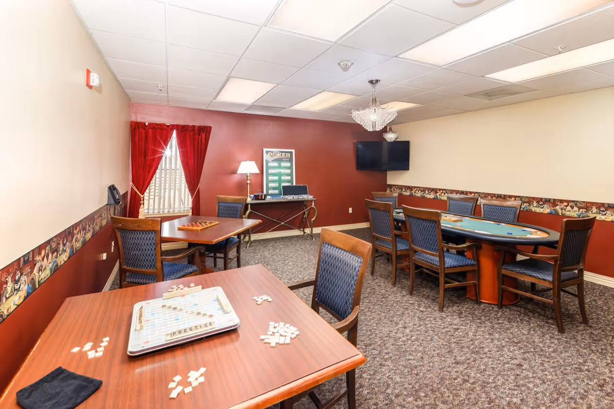 Interior game room with multiple tables and chairs, a Scrabble board in the foreground, a poker table, TV and red-accented walls.