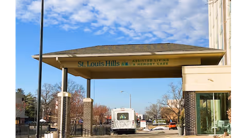 Entrance canopy of St. Louis Hills Assisted Living and Memory Care facility with a driveway underneath, a white vehicle parked, and trees and buildings in the background under a partly cloudy sky.