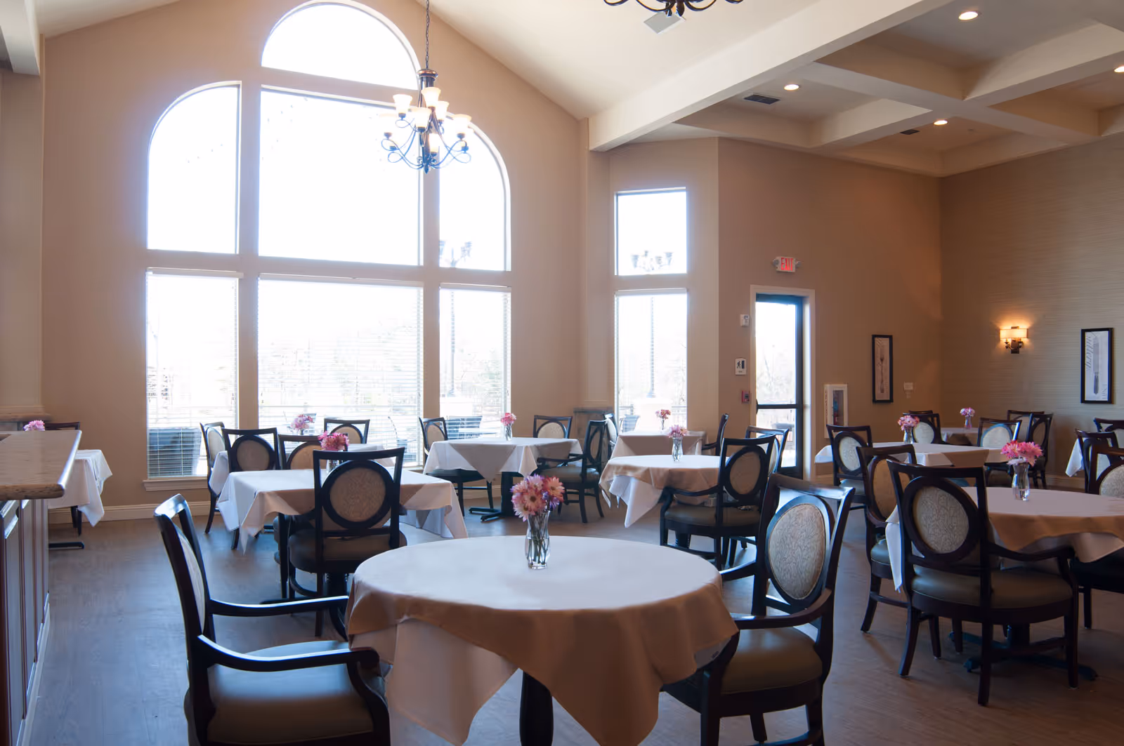 A bright dining room with round and rectangular tables covered in white tablecloths, chairs, large arched windows, and vases of pink flowers.
