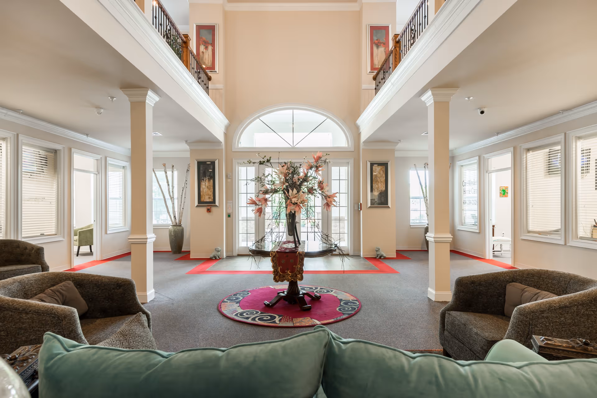 Spacious senior living facility lobby with high ceilings and large arched window above double glass doors. The room features a central round table with a floral arrangement, surrounded by comfortable armchairs and sofas. The walls are adorned with framed artwork, and there are large windows with blinds letting in natural light. The space has a warm and inviting atmosphere with beige walls and carpeted floors accented by red borders.