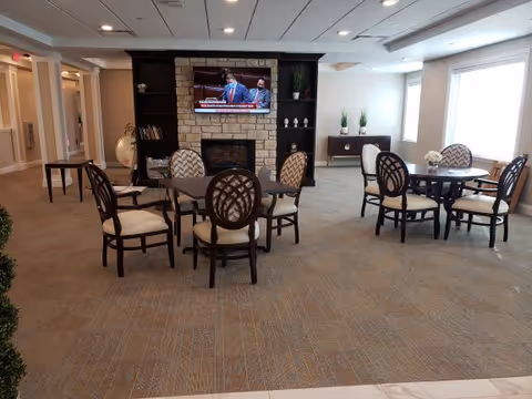 A senior living facility common area with multiple round tables and cushioned chairs arranged on a carpeted floor. A stone fireplace with a mounted flat-screen TV above it is visible in the background, flanked by built-in shelves with decorative items. Large windows on the right side allow natural light to fill the room.