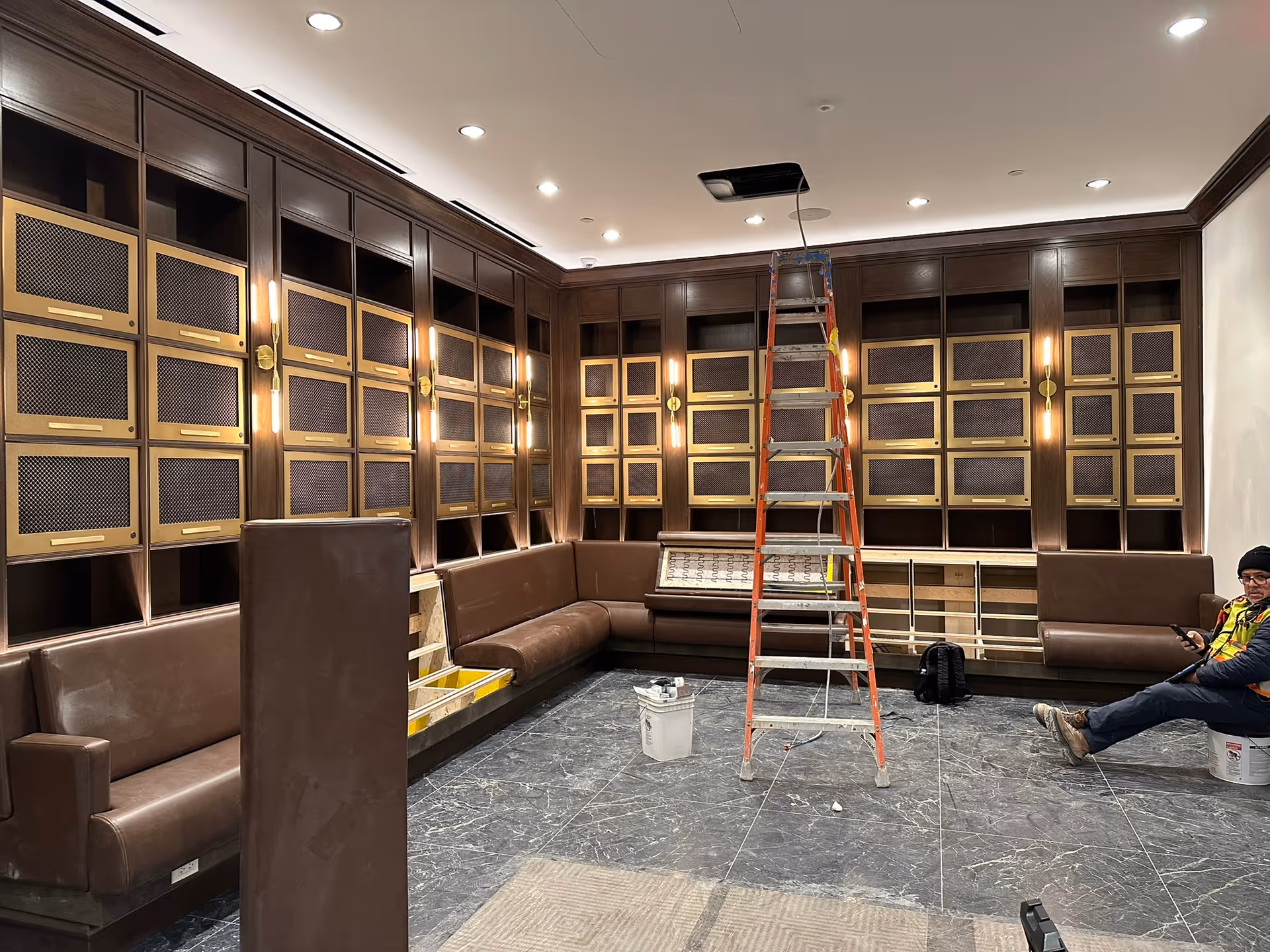 An interior room with dark wood paneling and built-in seating along the walls. The walls feature multiple small compartments with brass or gold-colored mesh doors. A red ladder is set up in the center of the room beneath an open ceiling panel. A worker in a safety vest and hat is sitting on a bucket on the right side, using a phone. The floor is covered with dark marble tiles, and there are some construction materials and tools scattered around.