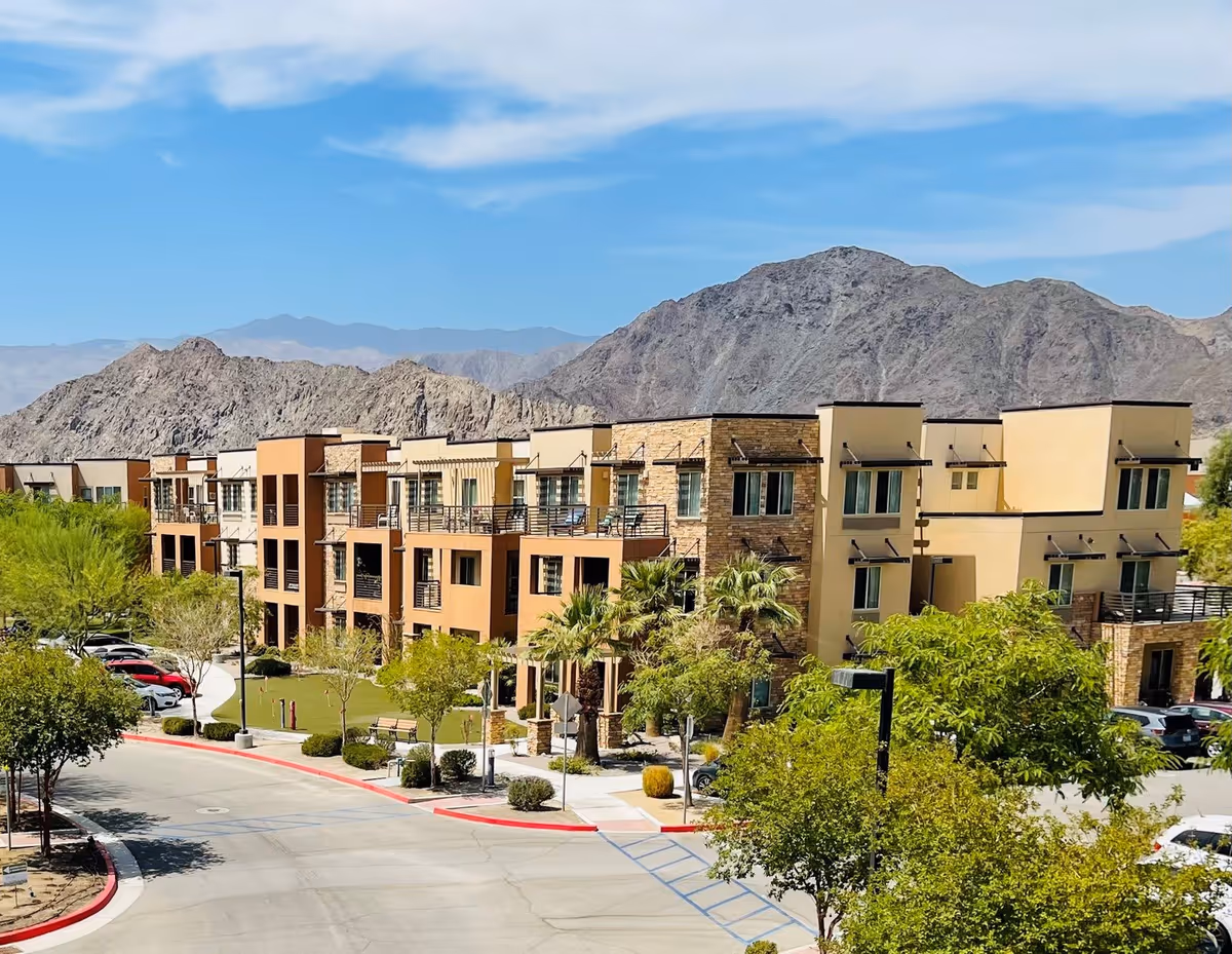 Exterior view of a modern multi-story senior living facility with balconies, surrounded by trees and parked cars, set against a backdrop of rocky mountains under a blue sky with scattered clouds.