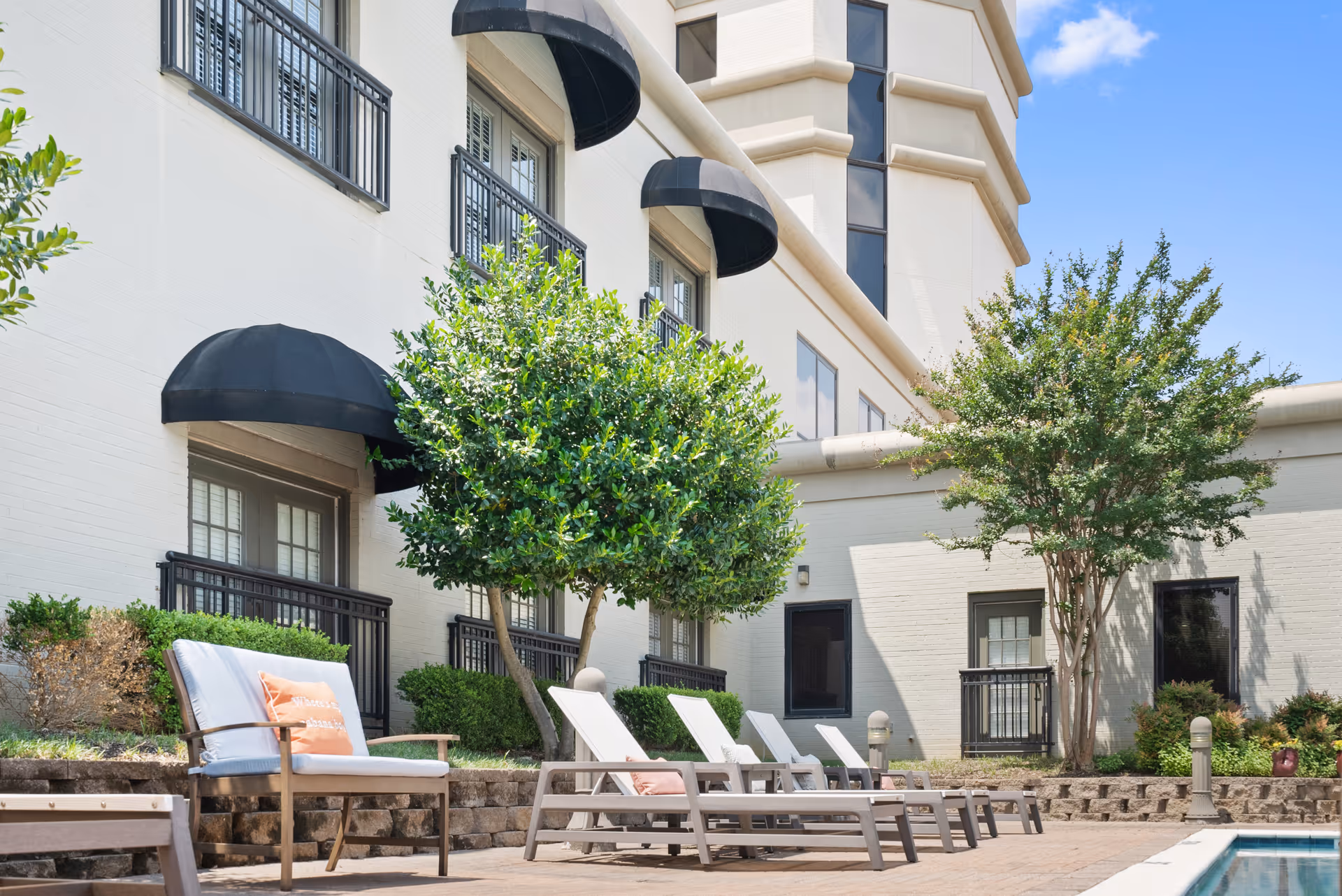 Outdoor patio area at Parc Place with several lounge chairs and a cushioned chair arranged near a swimming pool. The area is surrounded by a building with black awnings over windows and small trees and shrubs providing greenery.