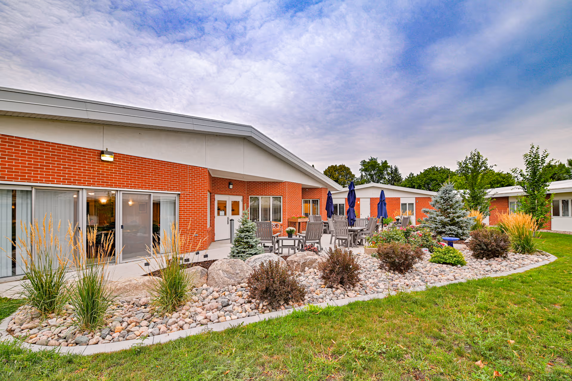 Outdoor patio area at Valley Senior Living on Cherry - Tufte Manor featuring a landscaped garden with rocks, shrubs, and small trees. There are several tables and chairs with closed blue umbrellas on a concrete patio adjacent to a red brick building under a partly cloudy sky.