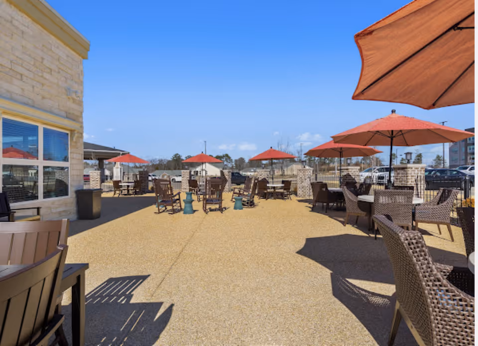 Outdoor patio area with multiple tables and chairs under red umbrellas on a sunny day, adjacent to a building with large windows.