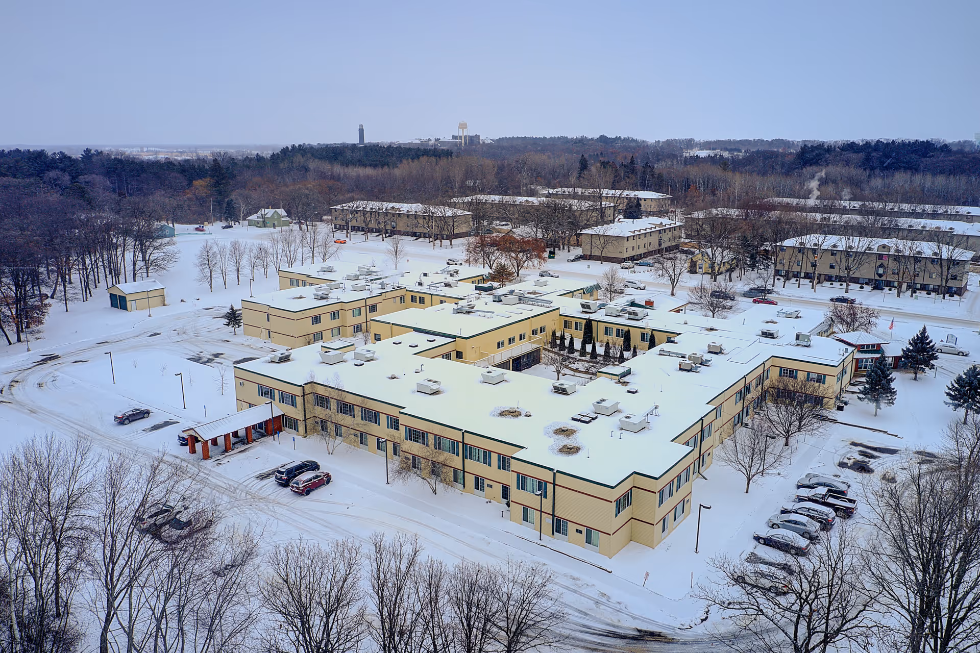 Aerial view of a large assisted living facility covered in snow during winter, surrounded by trees and parking lots with several cars. The building has a beige exterior with green trim and multiple sections connected in a U-shape.
