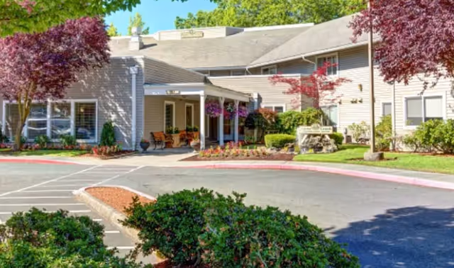 Front entrance of a senior living building with a covered driveway, benches, hanging flower baskets, and landscaped grounds.