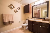 A bathroom with beige walls and floor tiles, featuring a toilet, a dark wood vanity with a granite countertop and a large mirror above it. There are two beige towels hanging on a towel rack on the left wall, and three decorative square wall hangings above the towels. The bathroom is well-lit with a light fixture above the mirror.