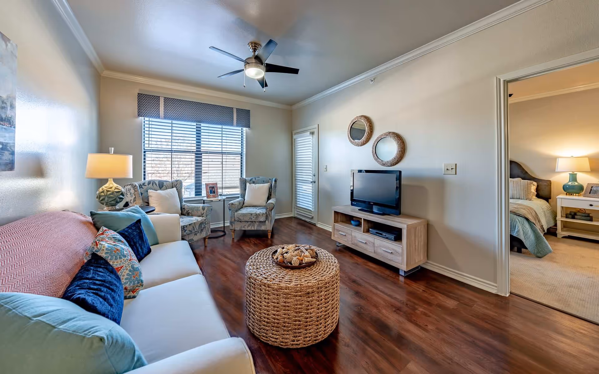 A cozy living room with a white sofa adorned with colorful pillows, two patterned armchairs near a window with blinds, a round woven coffee table in the center, a wooden TV stand with a flat-screen TV, and two round decorative mirrors on the wall. A doorway leads to a bedroom with a bed, nightstand, and lamp.