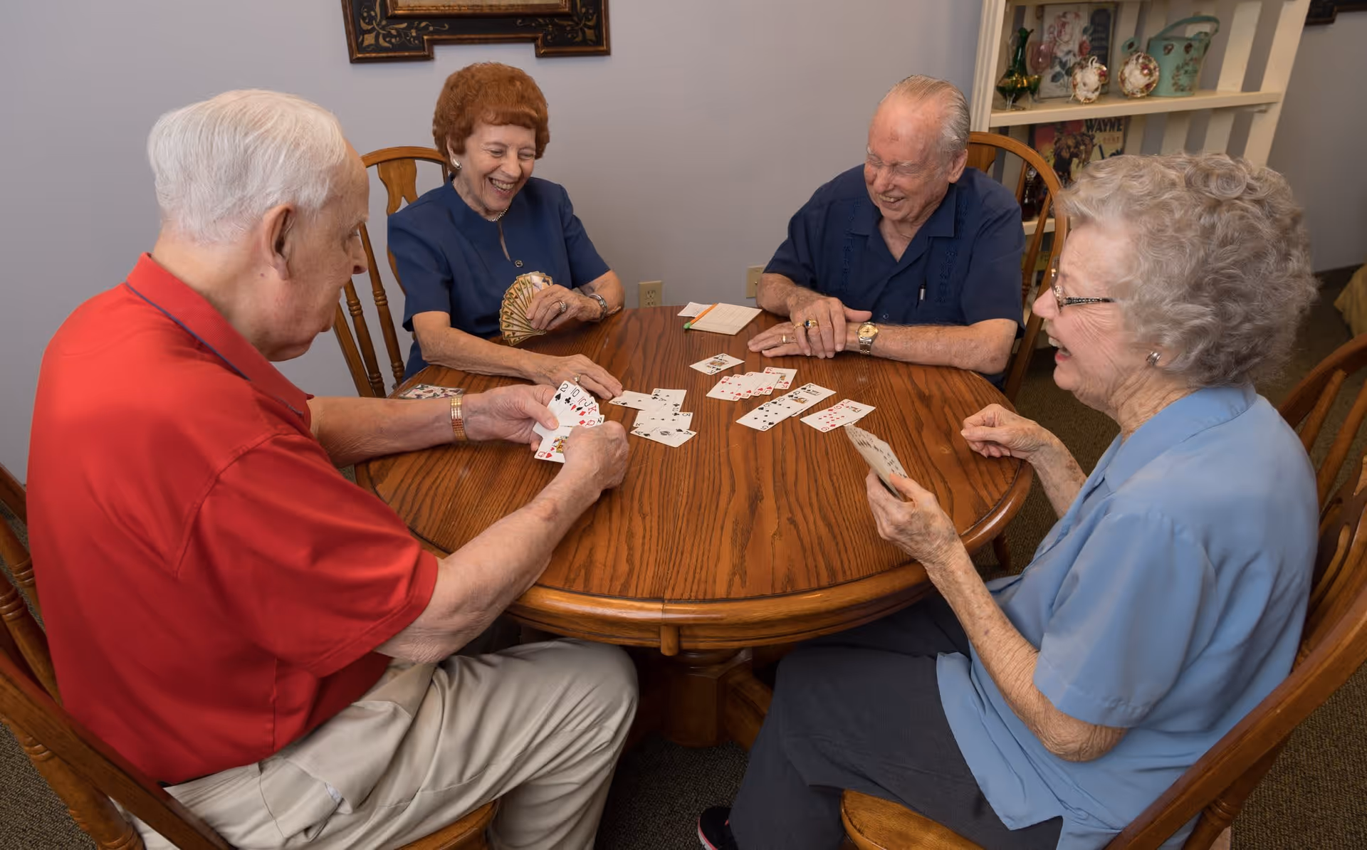 Four elderly people sitting around a wooden round table playing cards and smiling in a cozy indoor setting with shelves and decorative items in the background.
