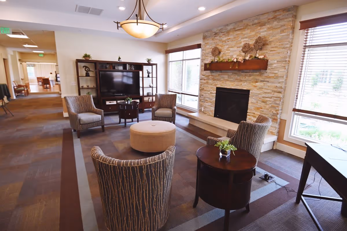 A cozy living room area in The Cortland Wyoming featuring four patterned armchairs arranged around a round beige ottoman. There is a dark wooden side table with a small plant, a large flat-screen TV on a wooden entertainment center, and a stone fireplace with a wooden mantel decorated with floral arrangements. Large windows with blinds allow natural light to fill the room.