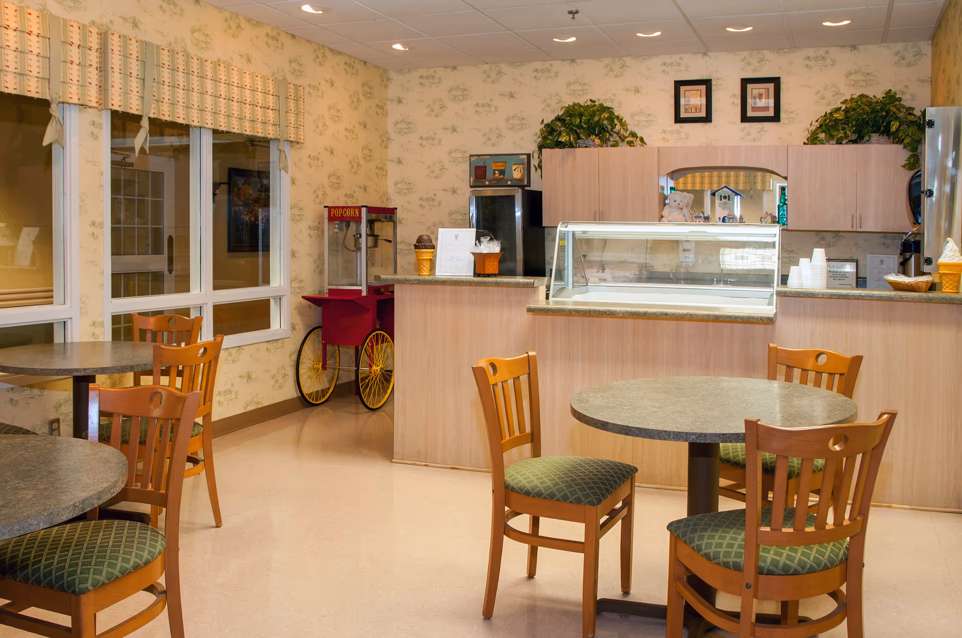 A small dining area with round tables and wooden chairs with green cushioned seats. In the background, there is a counter with a glass display case, a popcorn machine, and cabinets with plants on top. The walls have a light floral wallpaper and there are large windows with patterned valances.