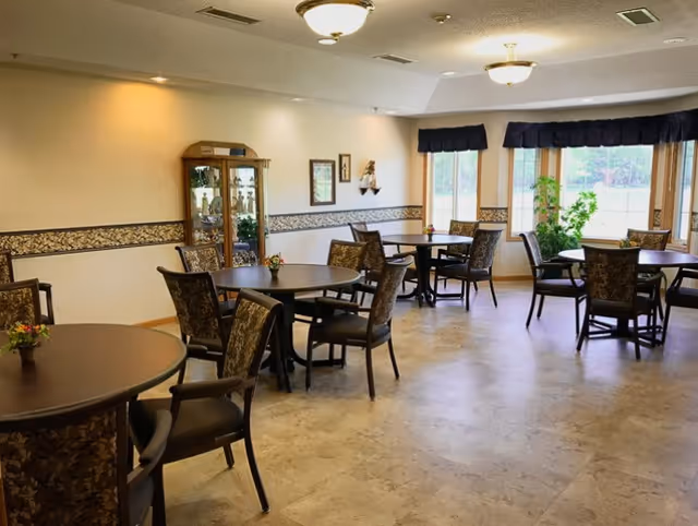 A bright dining room with several round tables and cushioned chairs arranged around them. The room has large windows with dark valances letting in natural light, a glass display cabinet in the corner, and decorative wall trim. Small flower arrangements are placed on the tables.