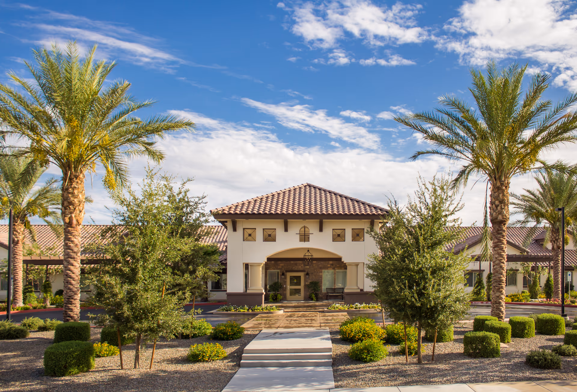 Front exterior view of a senior living facility with a tiled roof, surrounded by palm trees, shrubs, and a landscaped garden under a partly cloudy blue sky.