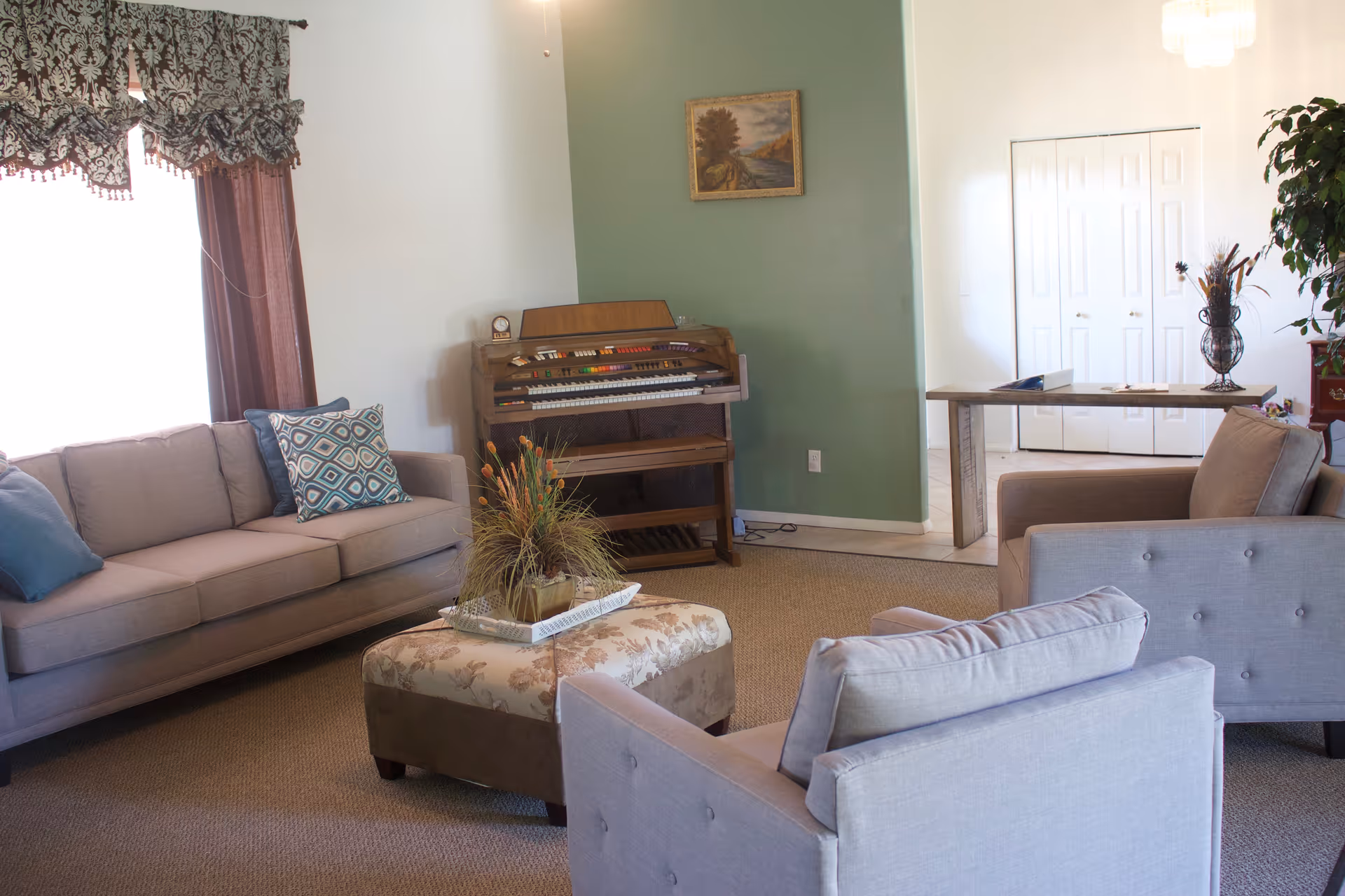 A cozy living room with a beige sofa, two matching armchairs, and a floral upholstered ottoman in the center. There is a wooden organ against a green accent wall with a framed painting above it. A window with patterned curtains lets in natural light, and a narrow table with decorative items is placed near a white double-door closet.