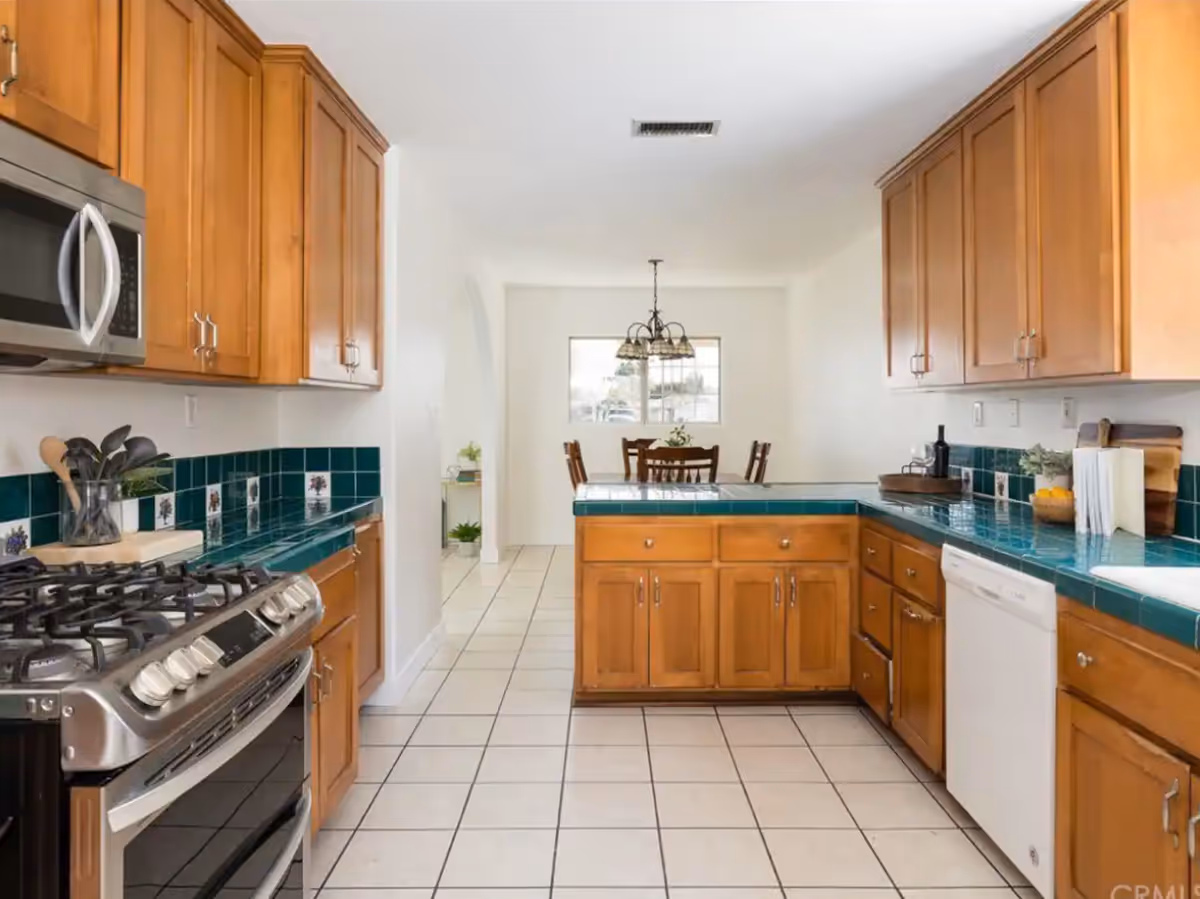 A bright kitchen with wooden cabinets, green tiled countertops, a stainless steel gas stove and microwave on the left, and a white dishwasher on the right. In the background, there is a dining area with a wooden table and chairs under a hanging light fixture.