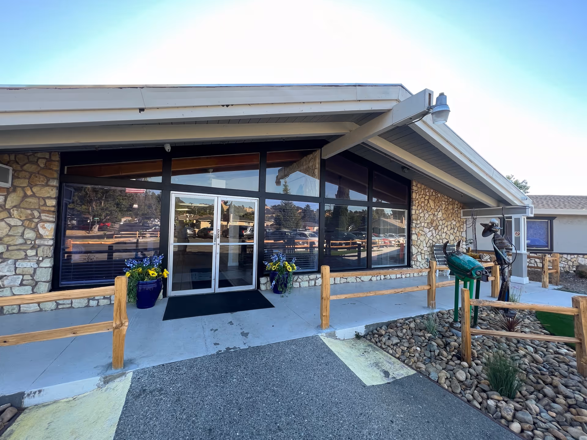 Front entrance of a single-story stone-faced building with large glass doors and windows, potted flowers, wooden railings, and a small metal sculpture.