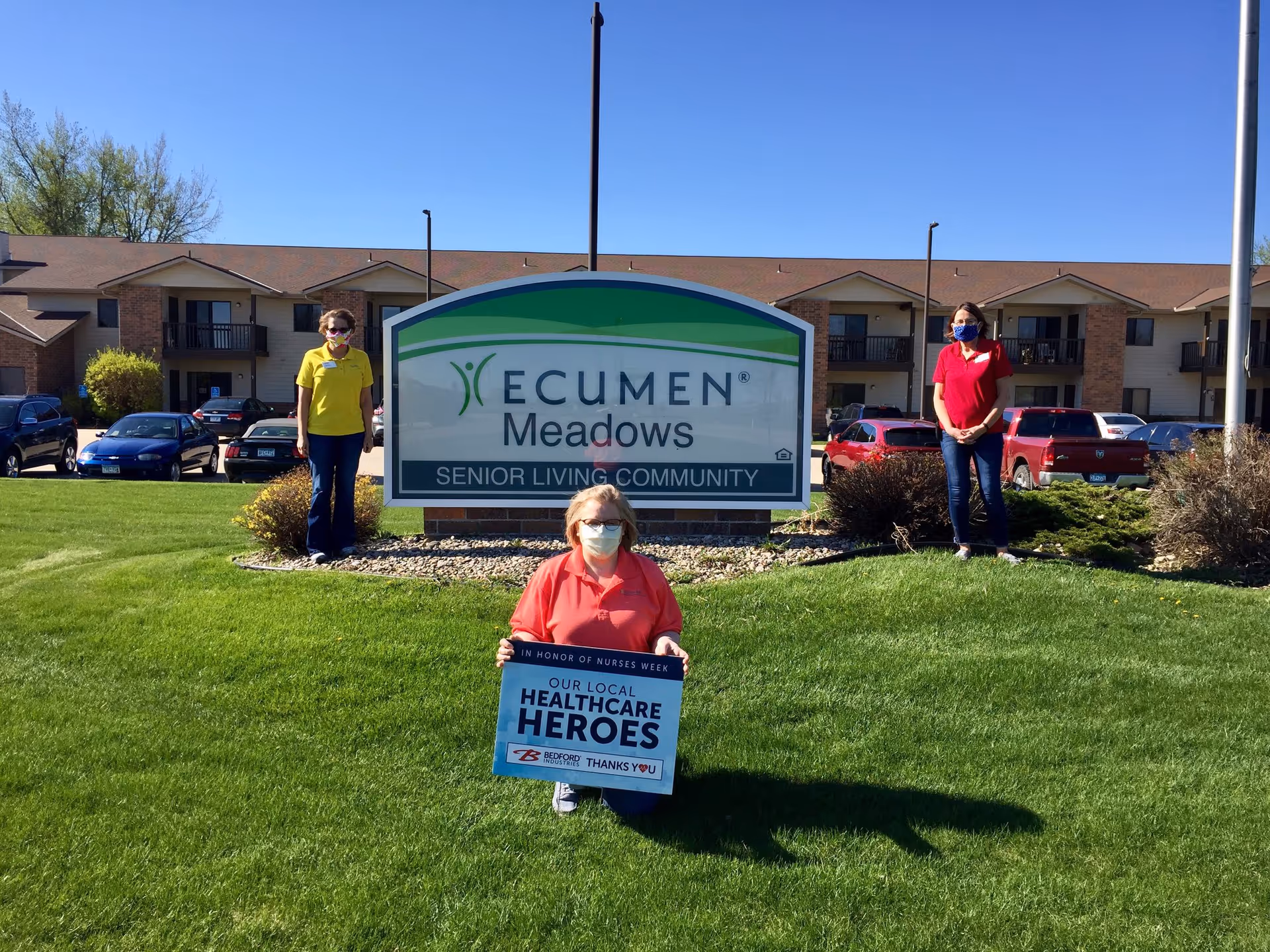 Three people wearing masks stand outside in front of a sign that reads 'Ecumen Meadows Senior Living Community'. One person in a red shirt is kneeling and holding a sign that says 'In honor of Nurses Week Our Local Healthcare Heroes'. The other two people stand on either side of the sign on a grassy area with a building and parked cars in the background under a clear blue sky.