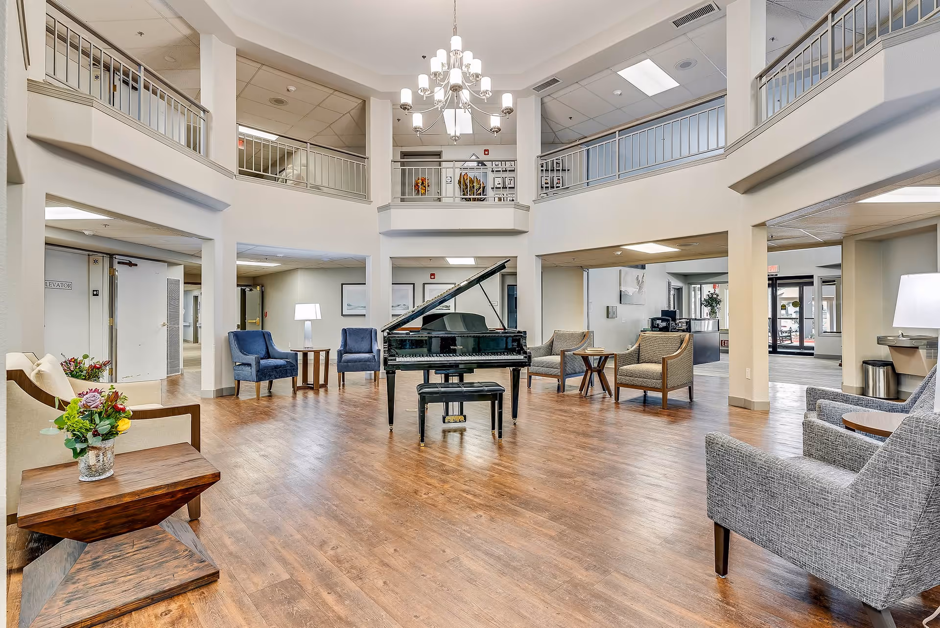 Spacious senior living facility common area with a grand piano in the center, surrounded by various seating options including blue and gray armchairs, wooden tables with flower arrangements, and a chandelier hanging from the ceiling. The area has a two-story open design with railings on the upper level and wood flooring throughout.