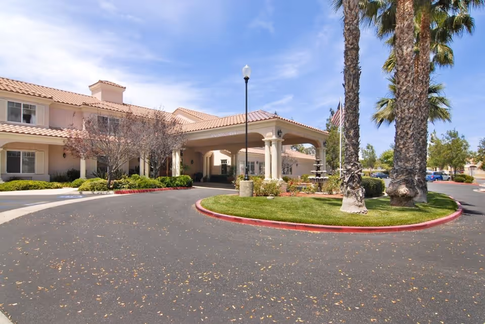 Front exterior of a senior living facility with a covered entrance supported by columns, palm trees, a fountain, and a circular driveway.