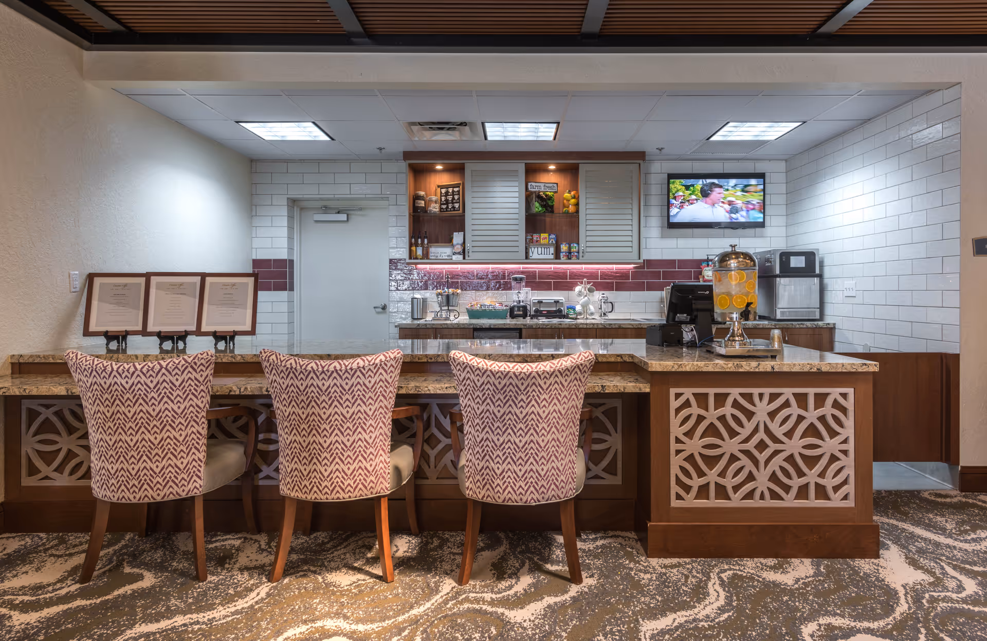A hospitality-style service counter with three patterned chairs, granite countertop, beverage dispenser, mounted TV, and tiled backsplash.