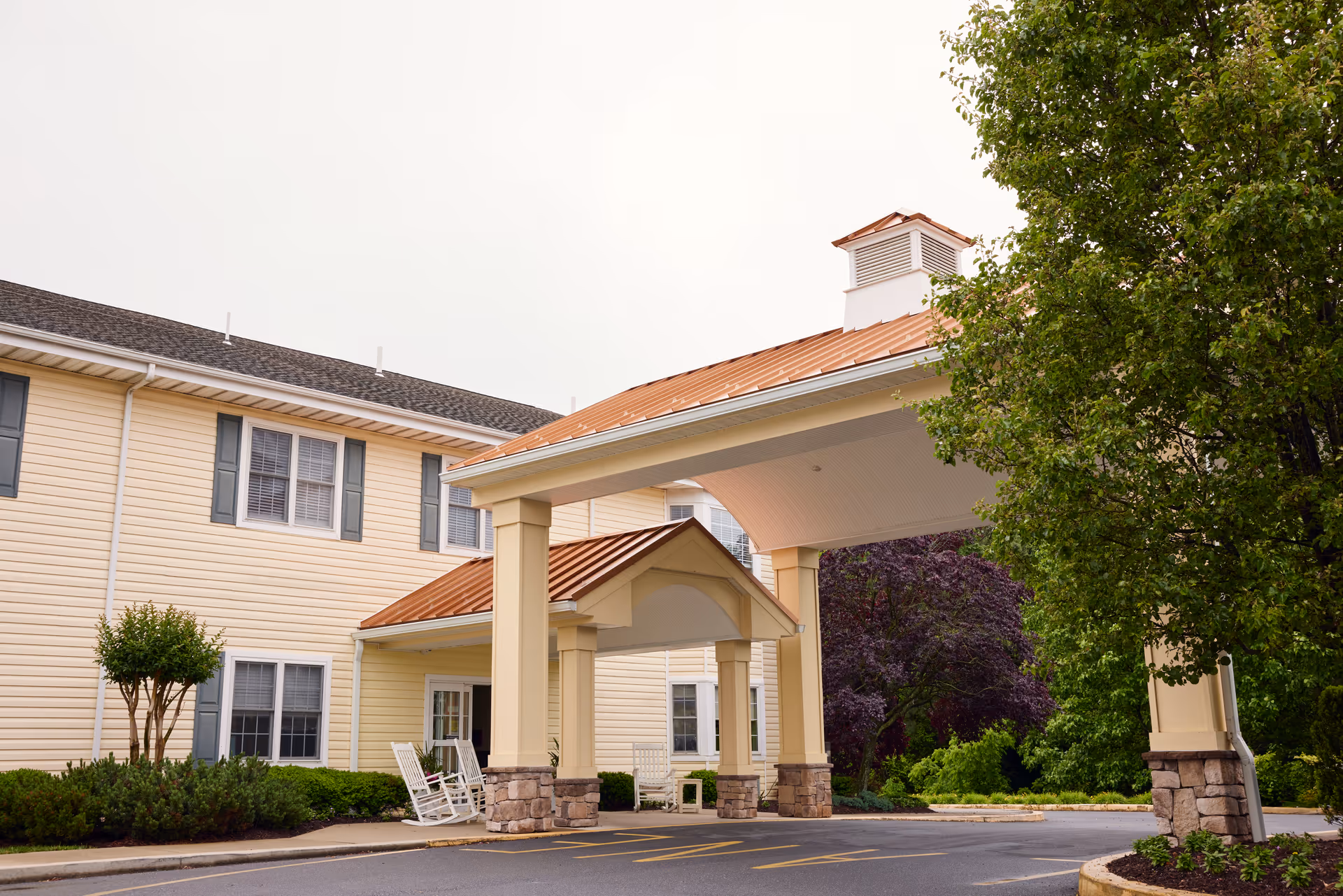 Entrance of a senior living facility with a covered drop-off area featuring a copper-colored roof supported by columns with stone bases. The building is light yellow with white trim and blue shutters. There are rocking chairs near the entrance and green trees and shrubs surrounding the area.