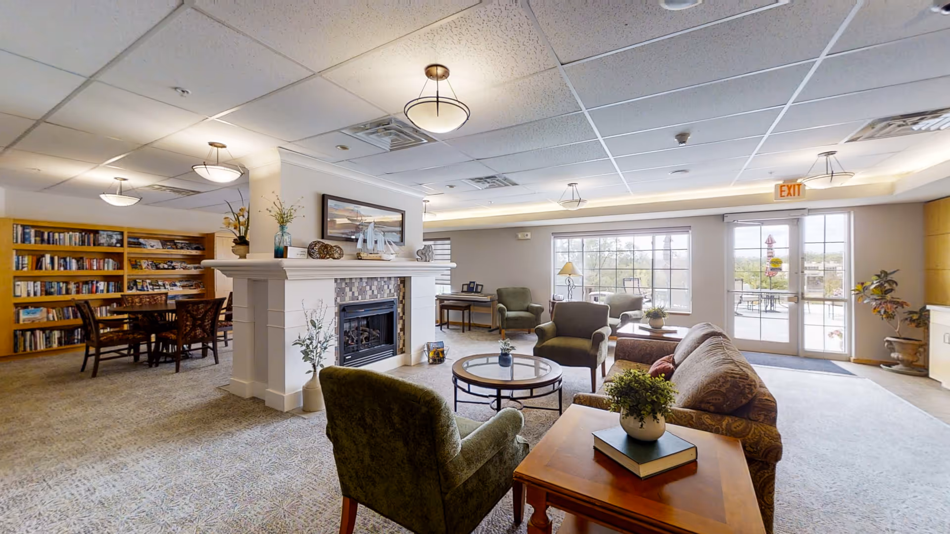 Bright communal living room in a senior facility with armchairs, a sofa, a central coffee table, a fireplace, bookshelves and large windows leading to an outdoor patio.